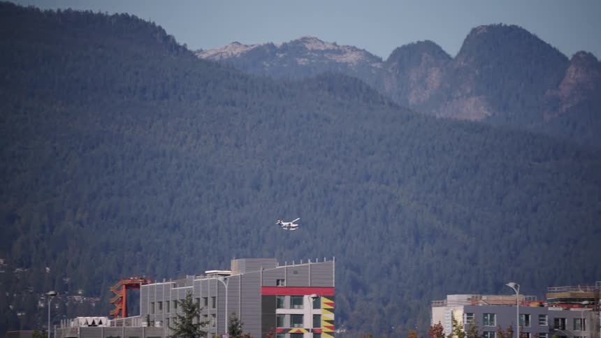 A small seaplane flies past city buildings and large industrial cranes with a dense tree covered mountain range in the background