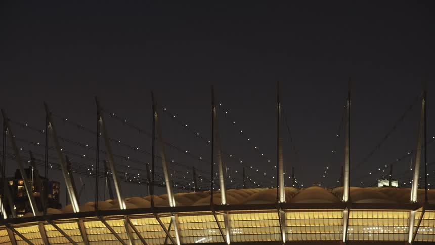 This wide lowlight shot captures the illuminated domed exterior of a large modern stadium or arena at nightthe structure is bordered by tall poles or cables receding into the dark sky creating a sense of scale and futuristic architecture