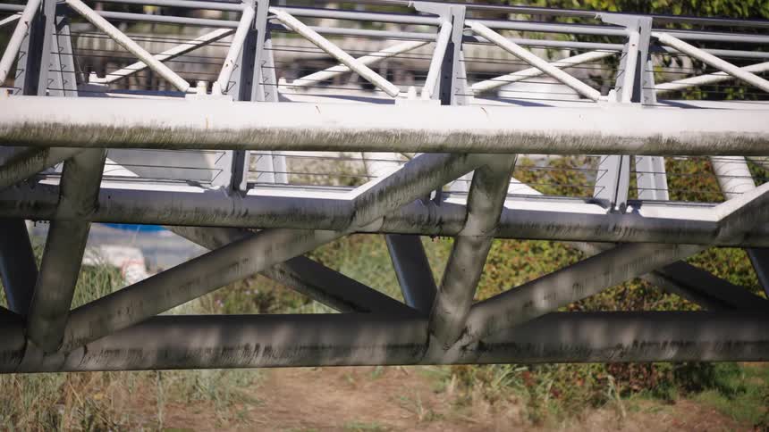 A metal pedestrian bridge with crisscrossed beams spans a green area as a couple of walkers and a dog pass overhead in bright afternoon sun