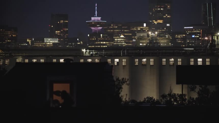 The person works in the dark room of the building in the foreground with a brigt city skyline in the background