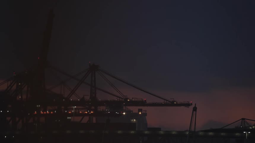 A cargo ports cranes loom against a deep night sky dotted with small lights and a faint orange glow on the horizon