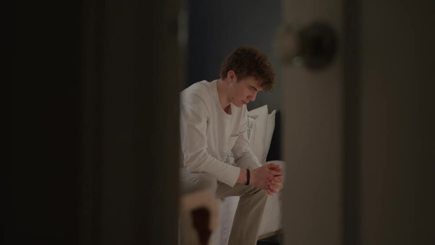 Medium view framed by a dark doorway of a man with lightcolored curly hair seated and looking down with his hands clasped wearing a white shirt and light pants in a dimly lit inner room