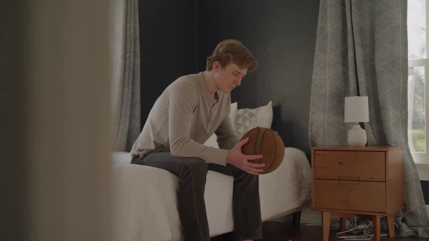 The teenager tosses the basketball while sitting on the bed