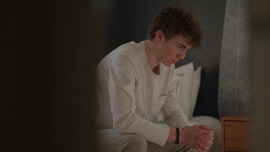 The teenager is sitting on the bed with his hands clasped