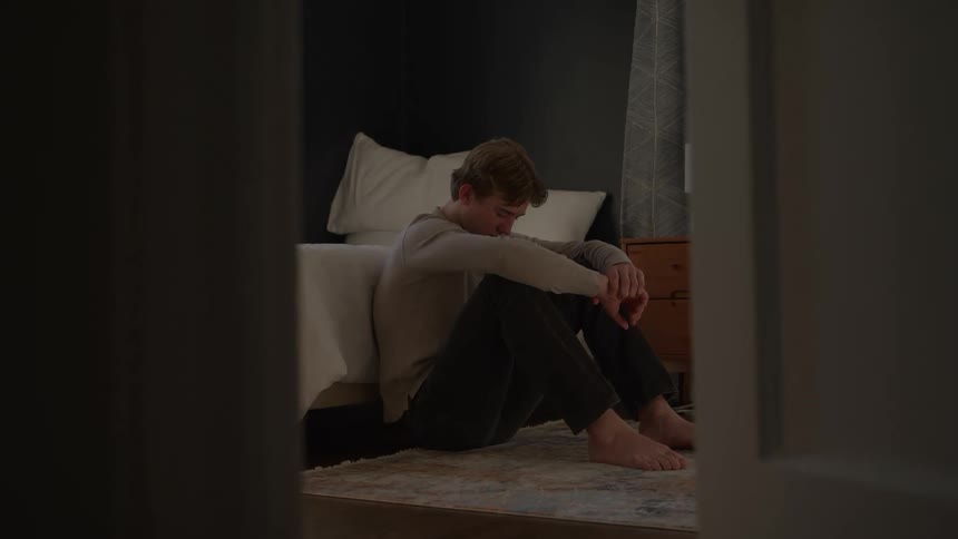 A young man sitting contemplatively on the floor beside a bed in a bedroom during daytime