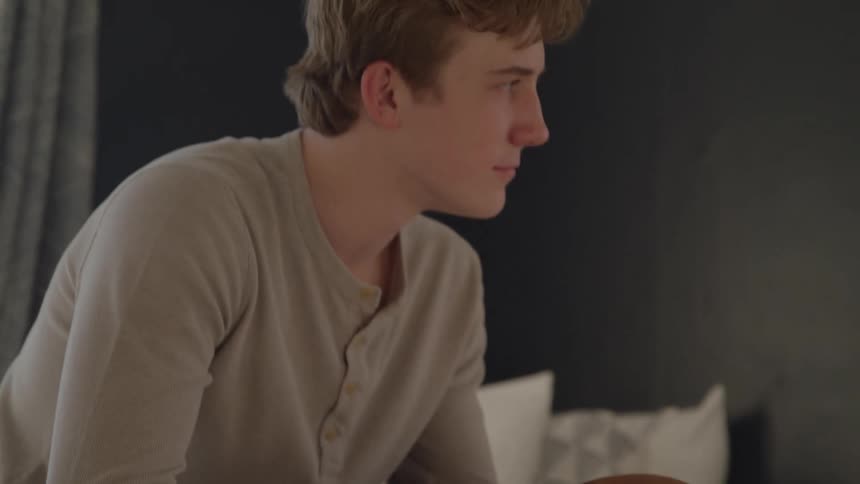 A young man sitting on the edge of a bed holding a basketball in his hands then slightly turning his head to look to the right with a serious expression
