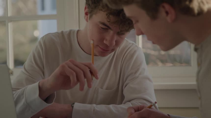 Two young male students collaborate intently on homework at a desk in a bright room working together with pencils and a laptop