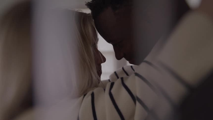 A close view shows a man and a woman with blonde hair in a striped shirt intimately embracing with their faces close together and a soft bright light surrounding them