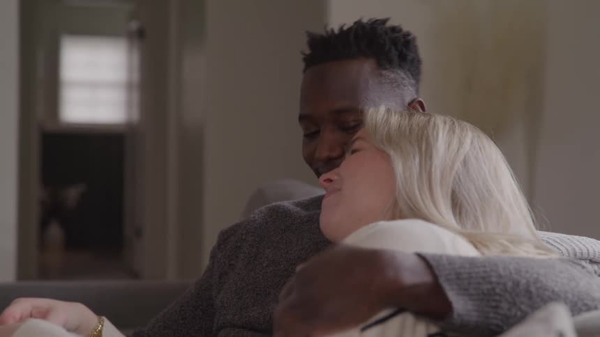 A smiling man with short dark hair wears a gray sweater and hugs a blonde woman while they sit close together on a couch watching television in a living room