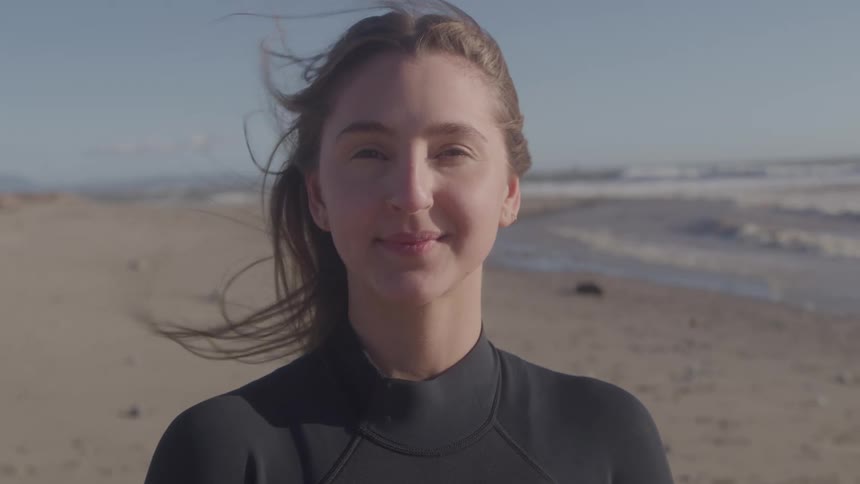 A smiling woman wearing a dark garment on a beach with wind blowing her hair looking straight ahead with a pale sand and distant ocean waves in the background