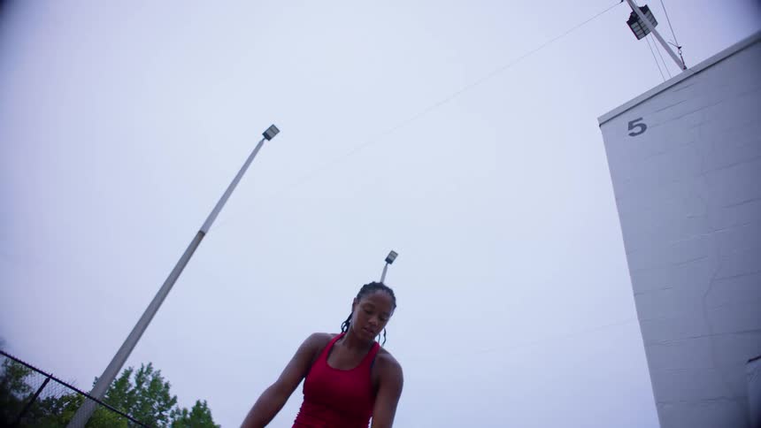 A lowangle view shows a woman wearing a red tank top as she throws a tennis ball into the air and swings a racket to serve on an outdoor court with a white wall and tall light poles visible against a bright overcast sky