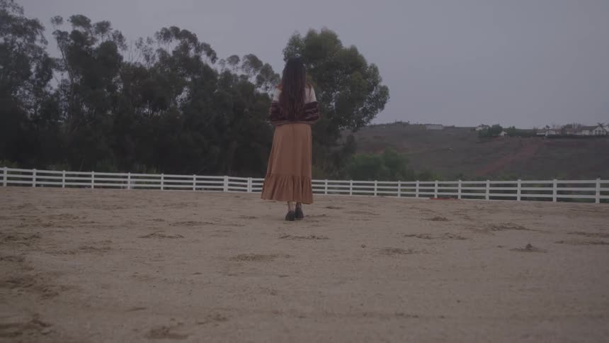 A woman walks across a dirt ground towards a white wooden fence line in an openair enclosure surrounded by trees and residential structures