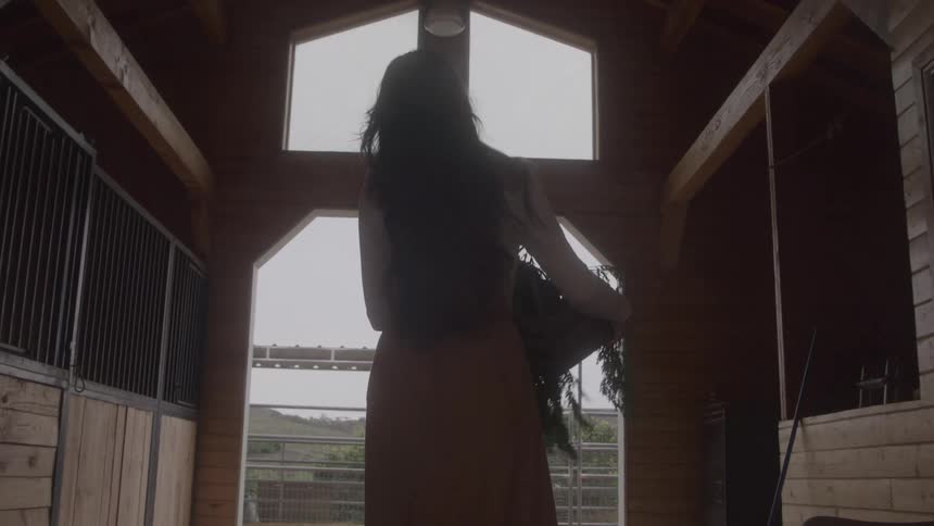 A young woman walks down a concrete aisle between wooden horse stalls carrying a crate of greenery and exits the barn structure toward a dirt enclosure