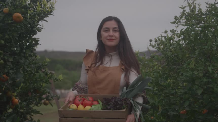 A woman standing between rows of fruit trees holding a wooden box filled with harvested vegetables and smiling directly at the camera