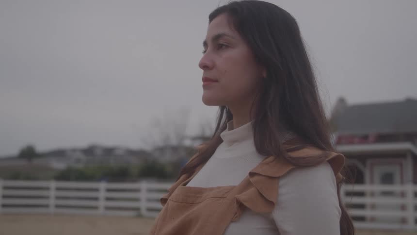 A woman with dark hair standing outdoors on a dirt area and looking toward the left with a white fence and a house visible in the softlit background