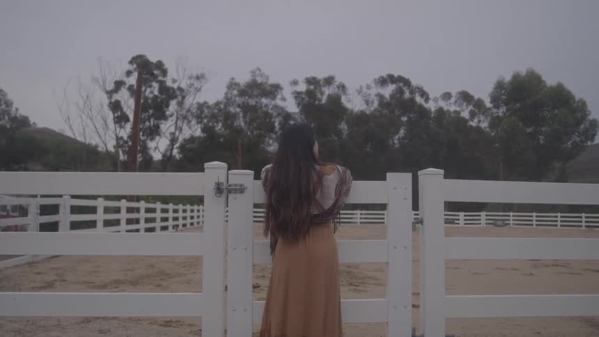 A woman with long dark hair wearing a tan dress and a shawl stands with her back to the camera looking over a white wooden fence into a horse corral with trees and a cloudy sky in the background