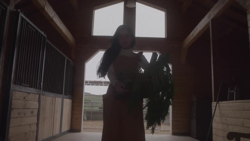A woman with long dark hair wearing a tan apron and a white shirt walks from a bright exterior into a wooden barn aisle holding a wooden crate overflowing with fresh green vegetables