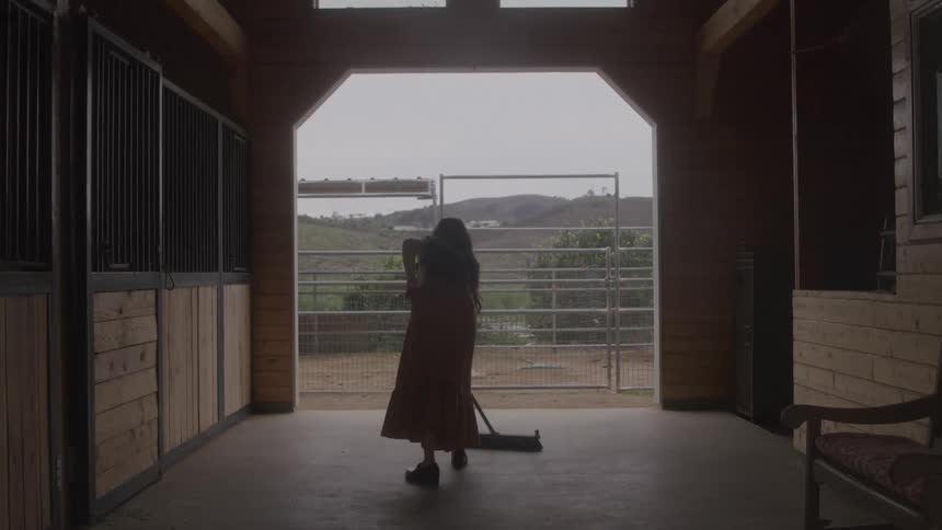 A woman is mostly silhouetted while sweeping the floor inside the central aisle of a barn and moves toward the open doorway facing an outdoor pasture and hills