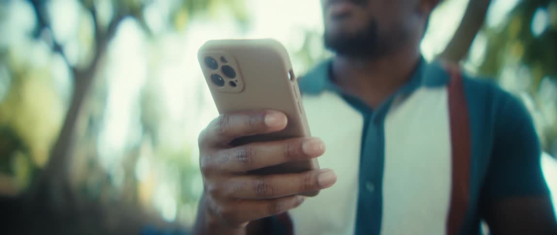 A young man holds a mobile phone with a brown case and uses the screen with his fingers while standing outdoors with trees in the background