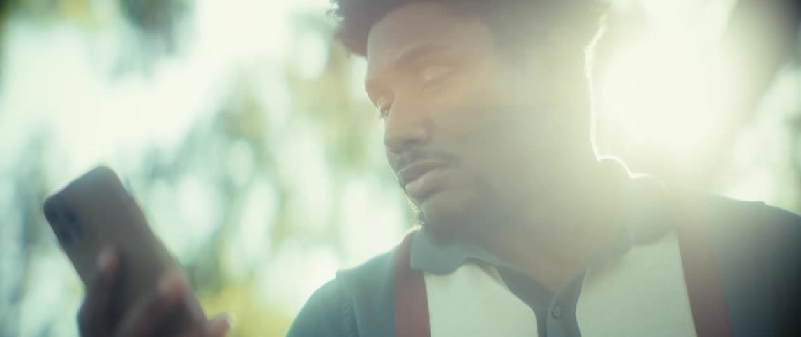 A close view shows a young man with curly hair wearing a teal and white polo shirt looking down and smiling while scrolling on a smartphone with a bright lens flare obscuring the frame outdoors in a sunny park