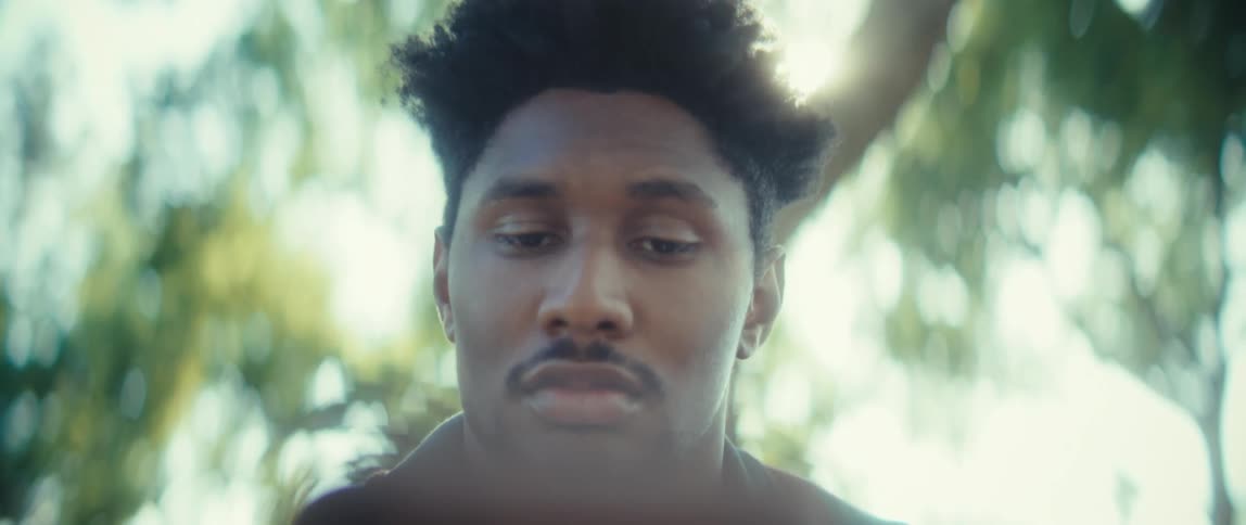 A focused young man looks down intently then slowly closes his eyes in contemplation against a brightly backlit blurry background