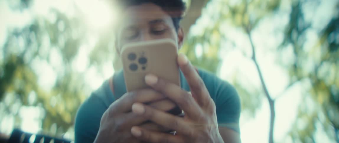 A young man wearing a teal and white polo shirt holds a smartphone with a tan case while scrolling and smiling outdoors in a park with bright sunlight filtering through the trees