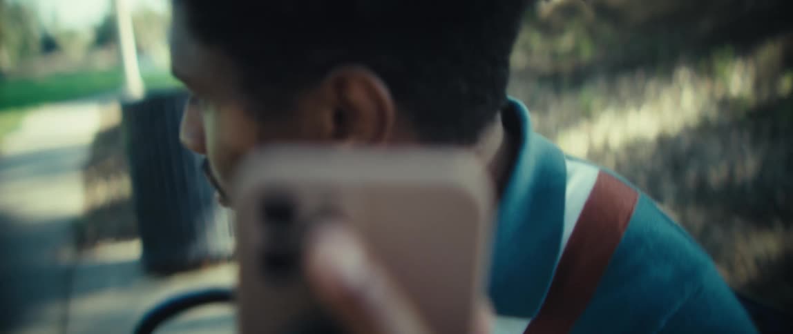 A closeup view focuses on the multiple camera lenses of a lightcolored smartphone as a man wearing a blue and red collared shirt brings the phone to his ear