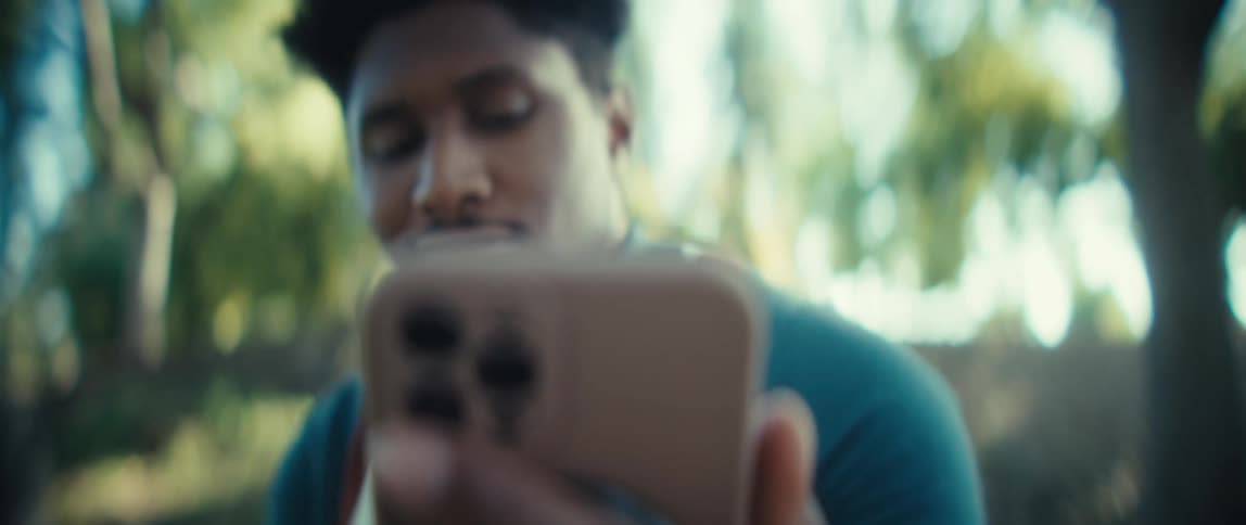 A young black man sits on a bench in a public park at midday holding his smartphone beneath the shade of a tree