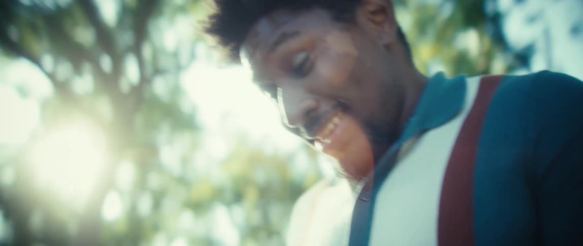 A close view shows a young black man with short dark hair wearing a teal polo shirt smiling widely while looking at and tapping a smartphone in a bright sundappled park