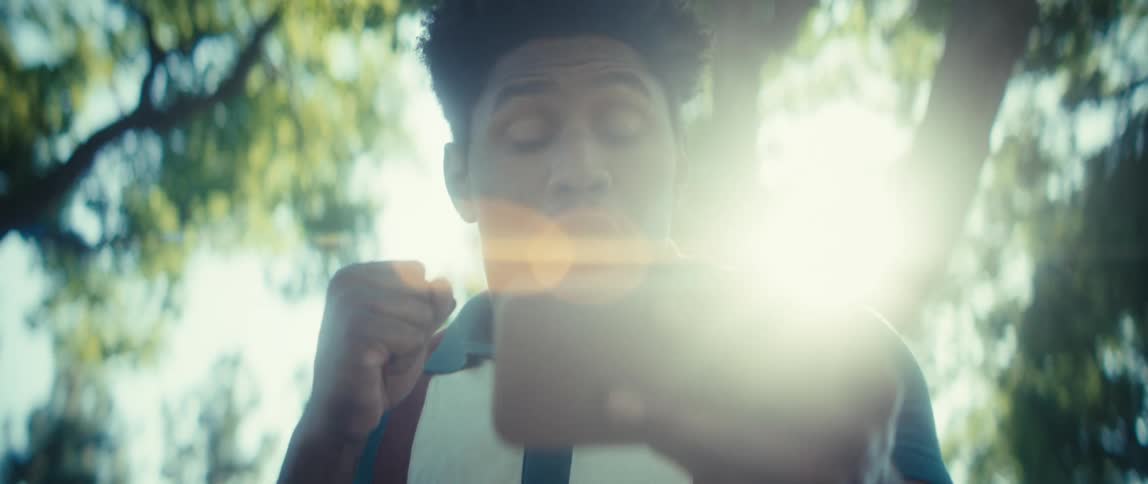 A young man wearing a teal and white polo shirt with brown suspenders holds a smartphone horizontally before reacting with a large joyful smile and a celebratory fist pump beneath the green canopy of sunlit trees