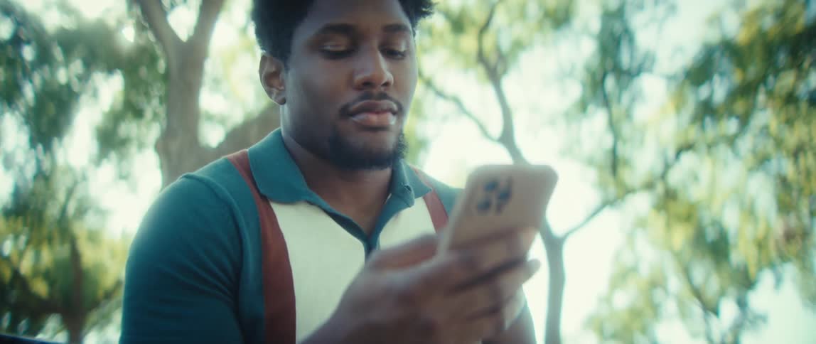 A young man sits on a bench under a tree on a sunny day scrolling through his phone with a smile on his face