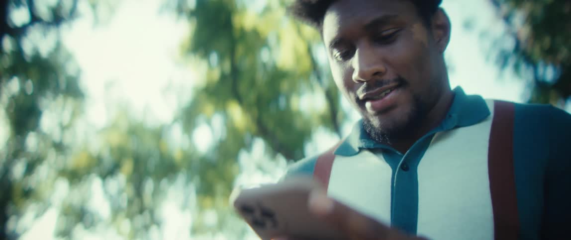 A young man smiles while scrolling on his phone with the sun creating a bright lens flare in a lush green outdoor setting
