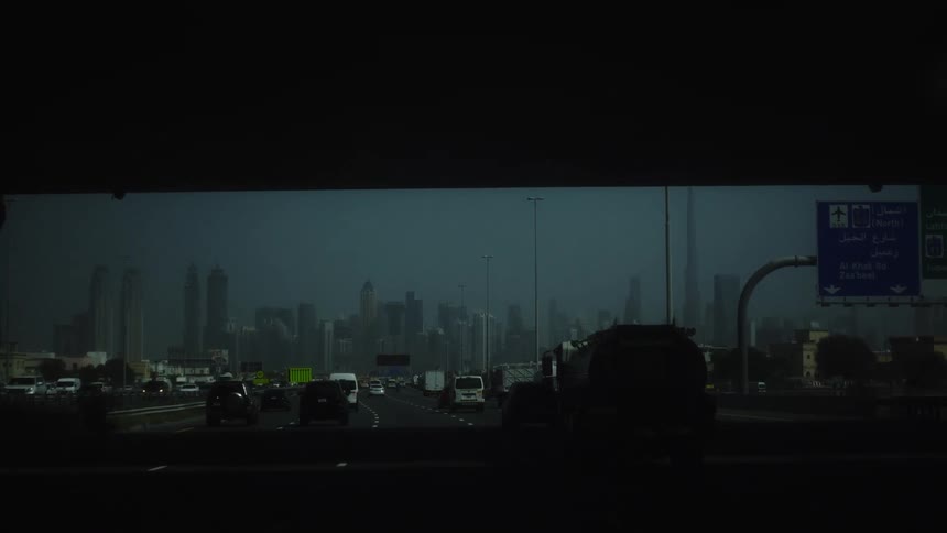 Traffic moving in both directions toward a dense distant collection of towering structures under a pale sky with directional signs visible on the right