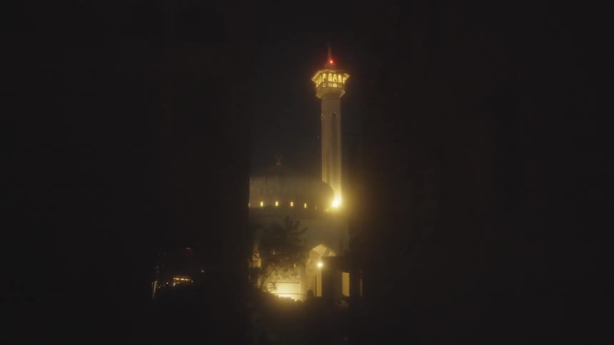 A tall narrow minaret with a red light at its peak is brightly illuminated by warm yellow light on a dark night seen through a silhouetted archway or trees