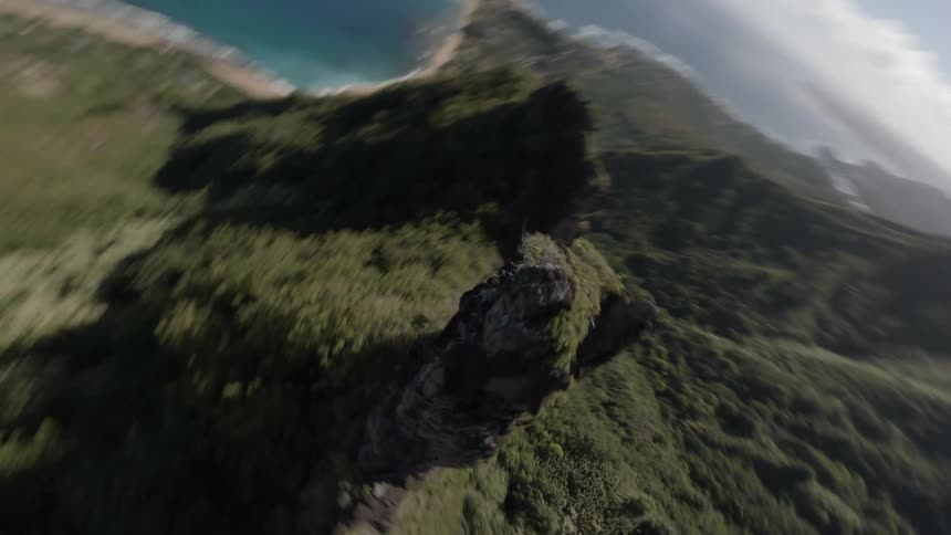 A fast aerial shot sweeps down and around a tall jagged rock spire covered in bright green moss and jungle vegetation with a blue ocean and coastline visible below