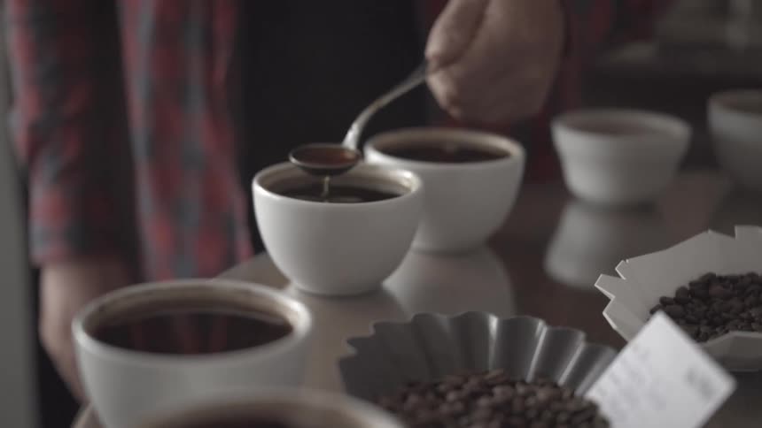 A man pouring coffee with a metal spoon into a coffee cup on a table indoors during daytime