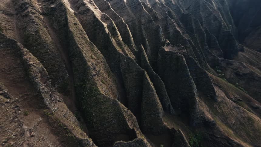 An aerial close view tracks down the steep deeply ridged slopes of a dark green and brown mountain range showing the dramatic textures and shadows of the terrain