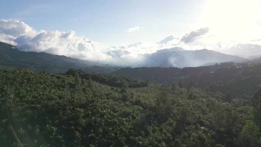 An aerial wide view shows a lush green mountain landscape with a small redroofed house and white clouds hugging the mountain peaks under a bright sunny sky