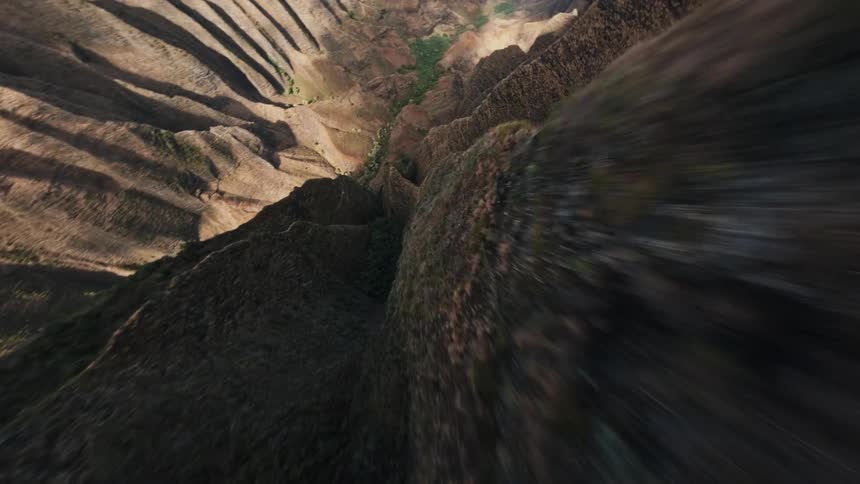 An extreme aerial drone view flies quickly along the sharp steep ridge of a rugged mountain valley with shadowed slopes leading down to the blue ocean and beach below