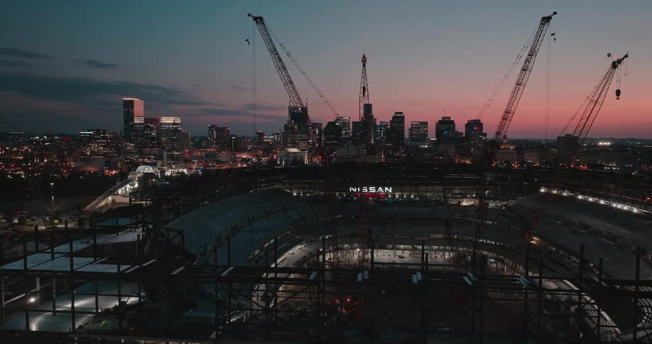 A stadium under construction at night with cranes positioned inside the structure and the city skyline glowing in the background under illuminated lights