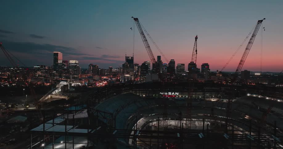 An aerial view shows a sports stadium under construction marked by several tower cranes with the skyline of a metropolitan city visible in the background against a darkening sky