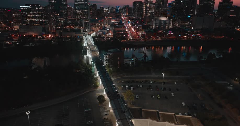An aerial camera moves backward and up over parking areas to reveal a city skyline illuminated by building lights and street lights with a long bridge crossing a dark river in the foreground during twilight