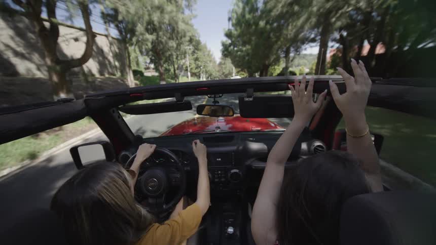Two young women cruise a red convertible with the top off hands raised feeling carefree and jubilant
