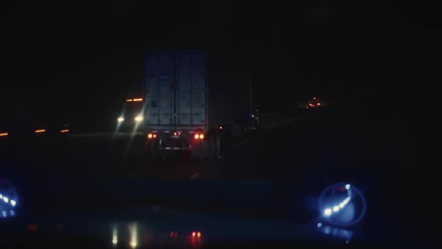 A semitrailer truck drives down a highway at night with the view framed by the bright illuminated side mirrors of the camera vehicle