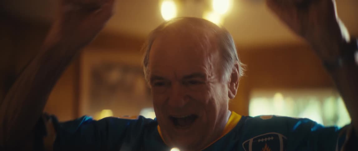 A medium closeup shot focuses on an older man wearing a blue and yellow sports jersey possibly a chargers jersey he is looking at a screen unseen and cheering enthusiastically pumping his fists and laughing suggesting a joyful moment wh