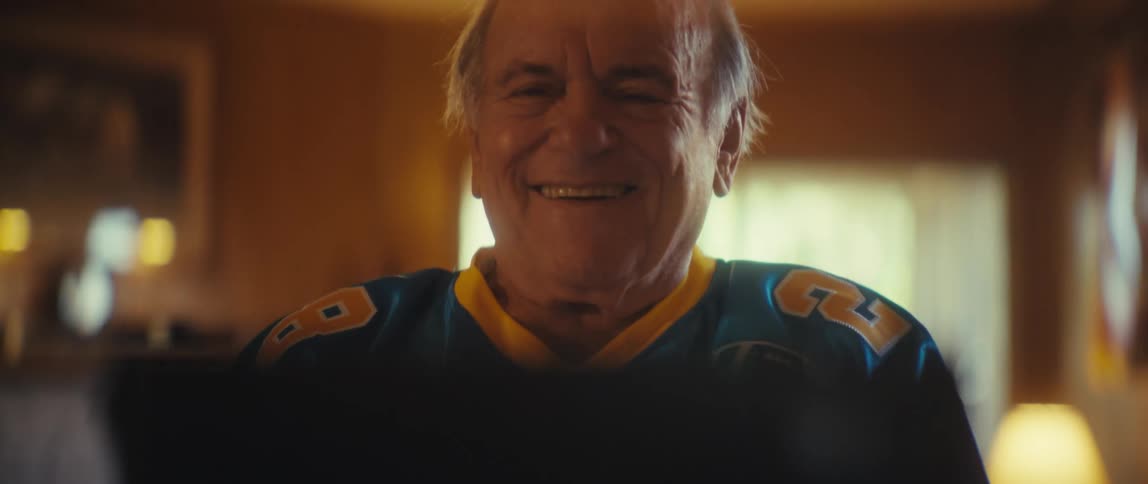 An elderly man in a blue football jersey beams with delighted surprise and clapping at a screen in his warmly lit living room
