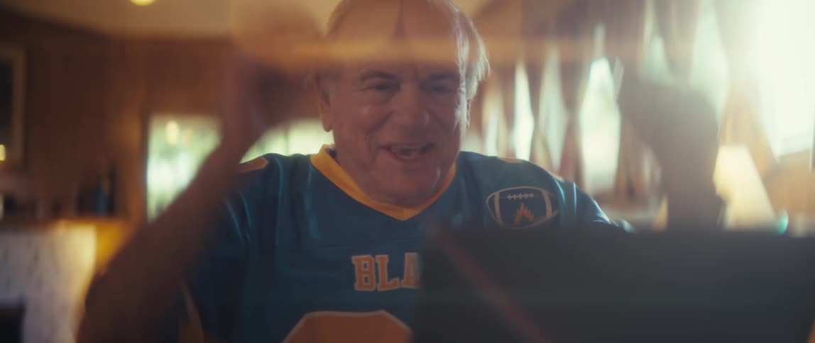 An elderly man sits in the living room wearing a blaze jersey watching a sports game on his mobile device with an excited expression