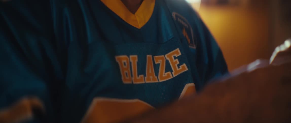 A closeup shot focuses on the chest of an older man wearing a blue and yellow sports jersey that reads blaze he looks down at a screen then looks up and bursts into genuine laughter expressing pure enjoyment
