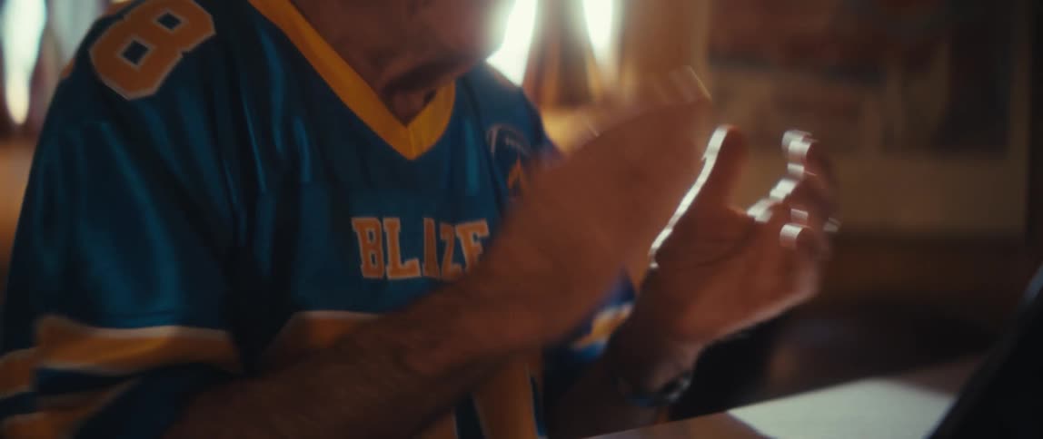 A bright sundrenched closeup shows an older man wearing a blue and yellow sports jersey he is actively gesturing with his hands and has an excited cheerful expression likely cheering on his team or reacting to an event on a screen
