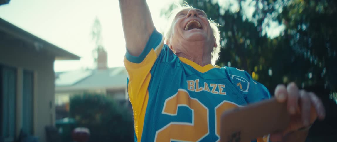 An elderly man stands outdoors at sunset wearing a jersey and watching a game on his phone suddenly bursting with excitement